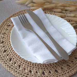 May include: A white plate with a white napkin, fork, and knife set on a beige crocheted placemat. Dried ornamental grass is in the background. The setting is for a meal.