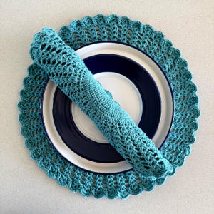 May include: A teal crochet placemat and rolled napkin sit atop a white plate with a navy blue rim and center. The placemat has a scalloped edge, and the napkin is rolled and placed on the plate. The items are on a light-colored surface.