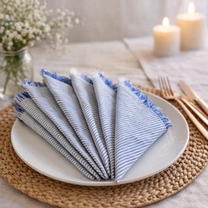 May include: Six folded white and blue striped napkins with blue fringing, arranged on a white plate. The napkins are on a woven placemat, with candles and cutlery in the background. The scene suggests a dining or table setting theme.