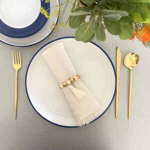 May include: A white plate with a navy blue rim, a gold fork, knife, and spoon, and a beige napkin with a beaded napkin ring and tassel. The table setting is arranged on a gray surface, with greenery in the background.