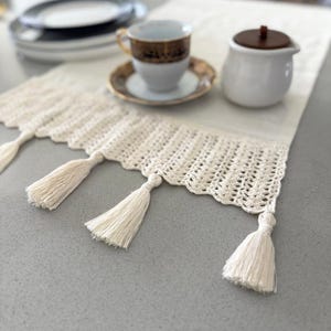 May include: Cream-colored table runner with a crocheted scalloped edge and tassels. A white teacup with gold trim sits on a saucer, alongside a sugar bowl and a stack of plates.