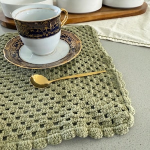 Könnte beinhalten: Eine weiße Teetasse mit goldenem Henkel und blau-goldenem Design steht auf einer passenden Untertasse. Ein goldener Löffel liegt auf einer salbeigrünen gehäkelten Tischmatte. Weitere weiße Teetassen sind im Hintergrund.