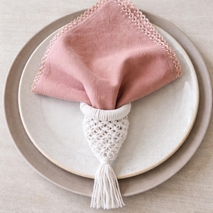 Può includere: Una tovagliolo di lino rosa antico con bordo all'uncinetto è tenuto da un portatovagliolo in macramè bianco con nappa. Il tovagliolo e l'anello poggiano su un piatto bianco, che si trova su un piatto grigio chiaro più grande.