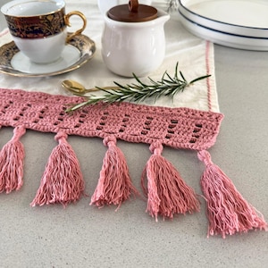 Pode incluir: Um corredor de mesa branco de crochê com uma borda de crochê rosa e borlas rosa. O corredor de mesa está sobre uma superfície cinza.