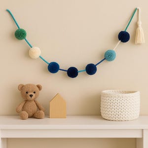Puede incluir: Una guirnalda decorativa con pompones azules, verdes y crema cuelga sobre una estantería blanca. Un oso de peluche de ganchillo, una casa de madera y una cesta blanca tejida están en la estantería. La guirnalda tiene una borla al final.