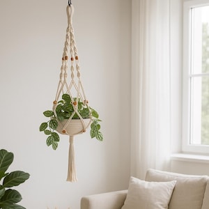 May include: A cream-colored macrame plant hanger with wooden beads, suspending a white ceramic pot filled with green leafy plants. The plant hanger is knotted and has a tassel at the bottom. The background includes a window and a neutral-toned sofa.