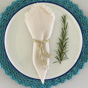 May include: A cream linen napkin with fringed edges is held by a gold and silver napkin ring with crystal accents. The napkin rests on a white plate with a blue rim, next to a sprig of rosemary. The plate sits on a teal crocheted placemat.