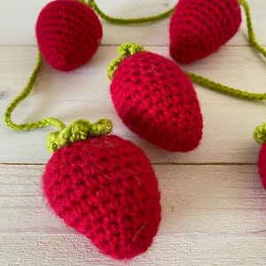 May include: A garland of crocheted red strawberries with green leaves. The strawberries are connected by a green yarn.