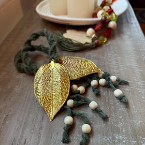 May include: Decorative gold leaf ornaments with intricate vein detailing. The leaves are attached to dark green macrame cords, accented with small, light-colored wooden beads. The leaves are approximately 4 inches long.