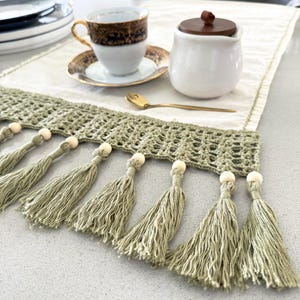 May include: A sage green crochet table runner with tassels and wooden beads. A white teacup with gold trim, a sugar bowl, and a gold spoon are on the table. The runner is approximately 30 cm wide.