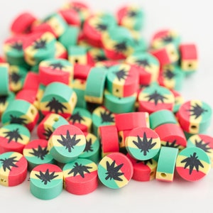 Puede incluir: Un montón de cuentas redondas de colores con un diseño verde, amarillo y rojo. Cada cuenta presenta un gráfico de hoja de marihuana negra.