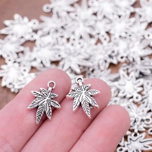 Puede incluir: Dos abalorios de hoja de marihuana plateados con un diseño detallado. Los abalorios se sostienen en una mano sobre un fondo de muchos abalorios similares.