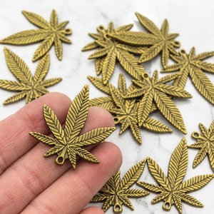 Puede incluir: Una colección de 12 abalorios de hoja de cannabis de metal dorado. Los abalorios están detallados con nervaduras de hojas realistas y tienen un agujero en la parte superior para colgar.