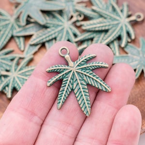 May include: A close-up of a hand holding a green and gold metal charm shaped like a marijuana leaf. The charm has a loop at the top for attaching to a necklace or bracelet.