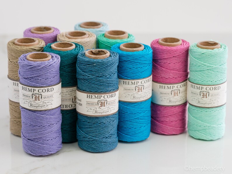 May include: Assortment of 10 spools of hemp cord in various colors, including lavender, teal, turquoise, pink, and mint green. Each spool has a label that reads "Hemp Cord" with a logo and the word "Hemptique".