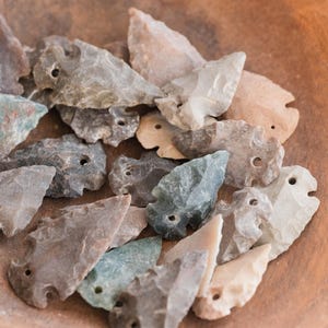 May include: A collection of stone arrowheads in various colors, including gray, tan, and teal, are displayed in a wooden bowl. Each arrowhead has a small hole near the top. The arrowheads vary in size and shape.