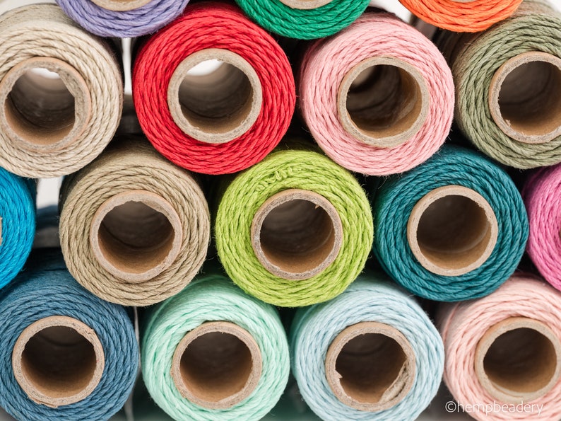 May include: Close-up of spools of colorful twine, including shades of green, blue, red, pink, and beige. The twine is wound around cardboard spools.