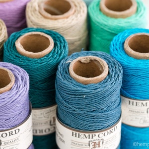 May include: Close-up of spools of hemp cord in various colors, including teal, blue, lavender, and natural. The spools are labeled "Hemp Cord" with a logo and the text "Premium Quality".