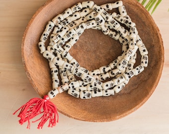 May include: A white prayer bead necklace with black markings and a red tassel. The beads are arranged in a circle on a wooden surface.