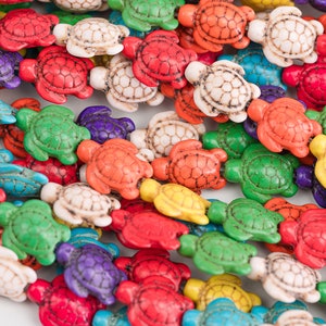 Op de afbeelding: Een streng met kleurrijke schildpadvormige kralen gemaakt van turquoise, rood, groen, wit, geel en paars. De kralen zijn aan elkaar geregen op een doorzichtige draad.