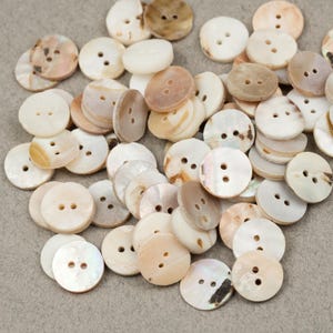 May include: A collection of round buttons in various shades of white, cream, and brown. The buttons have two holes and a slightly iridescent sheen. Some buttons have a marbled appearance, while others are solid in color. The buttons are scattered across a neutral surface.