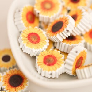May include: A collection of small, wooden sunflower-shaped embellishments. Each piece features a yellow and orange floral design with a dark brown center and white, scalloped edges. These decorative elements are ideal for craft projects.