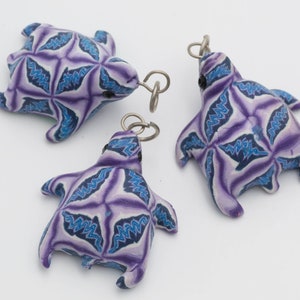 Puede incluir: Tres encantos de tortuga púrpura y azul con un patrón geométrico. Las tortugas están hechas de arcilla polimérica y tienen anillos de salto plateados.