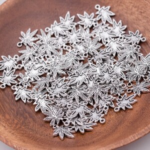May include: A large quantity of silver-toned metal charms in the shape of maple leaves. The charms have a detailed design and are scattered on a wooden surface.