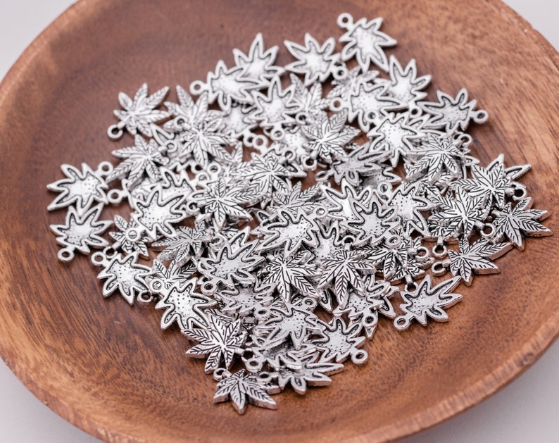 May include: A large quantity of silver-toned metal charms in the shape of maple leaves. The charms have a detailed design and are scattered on a wooden surface.
