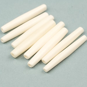 Op de afbeelding: Een verzameling van acht crèmekleurige, cilindrische kralen met een licht taps toelopend ontwerp. De kralen liggen op een lichtblauwe ondergrond en tonen hun gladde, gepolijste textuur. Geschikt voor sieraden maken en knutselprojecten, ongeveer 2 cm lang.