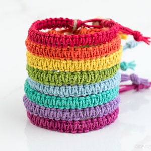 Puede incluir: Una pila de siete pulseras de macramé en un arcoíris de colores: rojo, naranja, amarillo, verde, azul, morado y magenta. Cada pulsera está hecha de cordón de cáñamo tejido y tiene un cierre ajustable. Las pulseras están apiladas una encima de otra.