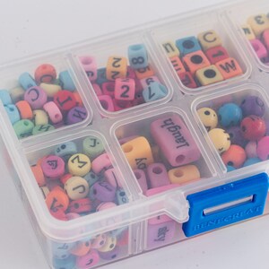 Puede incluir: Una caja de almacenamiento de pl&aacute;stico transparente con un cierre azul, llena de cuentas de letras y n&uacute;meros de colores. Las cuentas son de diferentes colores, incluyendo rosa, azul, amarillo, verde y morado. La caja tiene varios compartimentos, cada uno lleno de un tipo diferente de cuenta.