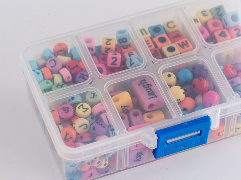 Puede incluir: Una caja de almacenamiento de pl&aacute;stico transparente con un cierre azul, llena de cuentas de letras y n&uacute;meros de colores. Las cuentas son de diferentes colores, incluyendo rosa, azul, amarillo, verde y morado. La caja tiene varios compartimentos, cada uno lleno de un tipo diferente de cuenta.