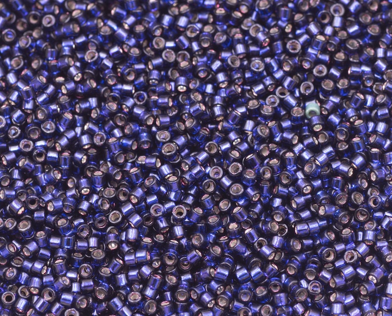 May include: A close-up shot of a pile of small, cylindrical beads. The beads are a deep, shimmering blue with a metallic sheen. The beads are scattered, creating a textured surface. The beads are likely used for jewelry making or other crafts.