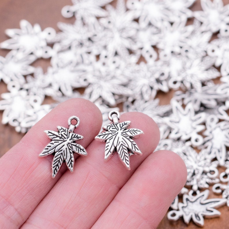 May include: Two silver-toned marijuana leaf charms with a detailed design. The charms are held in a hand against a background of many similar charms.