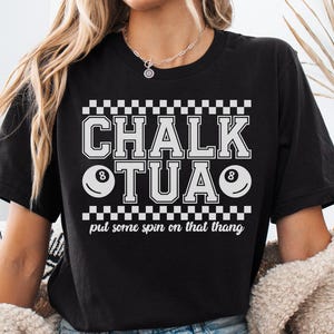 May include: Black t-shirt with the text "CHALK TUA" in a white, blocky font, with two eight-ball graphics. The phrase "put some spin on that thang" is below the main text. The shirt has a checkerboard design.