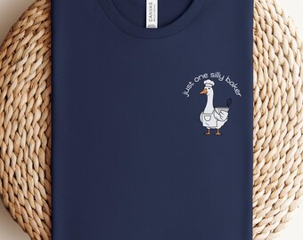 May include: Navy blue t-shirt with a round neck. The shirt features a small graphic of a goose wearing a chef's hat and apron, with the text "just one silly baker" in a curved font. The shirt is folded on a woven surface.