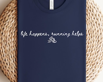 May include: Navy blue t-shirt with the phrase "life happens, running helps" in white script, along with a small running shoe graphic. The shirt is folded and rests on a woven, circular surface.