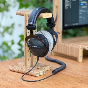 May include: Black headphones with a gray earpad on a wooden stand. The headphones have the text "DT 770 PRO" on the side. The stand is made of plywood and has a coiled cord.