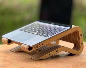 May include: A wooden laptop stand with a natural finish. The stand is designed with a curved, layered construction and features a built-in ledge to hold a laptop. The laptop is silver and black.