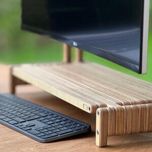 May include: A wooden monitor stand with a layered plywood design. The stand is raised off the surface and has a keyboard sitting in front of it.