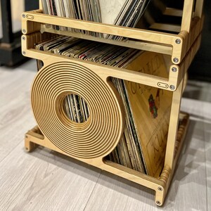 Puede incluir: Una unidad de almacenamiento de discos de vinilo de madera con un diseño de corte circular. La unidad está llena de discos de vinilo y tiene un acabado de madera natural.
