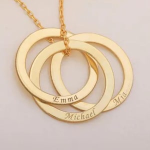Peut inclure: Collier doré avec trois anneaux entrelacés, chacun gravé d'un nom : Emma, Michael et Mia. Le pendentif est suspendu à une chaîne délicate. Le fond est un tissu blanc cassé.