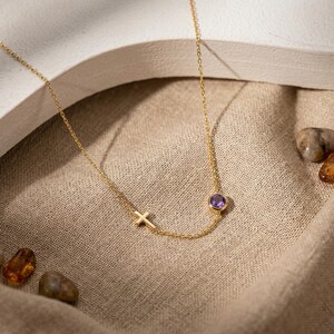 May include: A gold-colored necklace with a small cross pendant and a round, purple gemstone. The necklace is on a beige fabric surface, with small stones and amber-colored pieces scattered around.