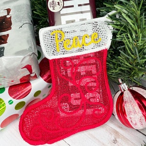 Puede incluir: Una media de Navidad roja y blanca con la palabra "Peace" bordada en amarillo en el puño. La media está llena de una barra de chocolate.