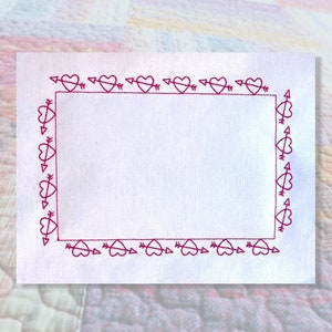 Könnte beinhalten: Ein weißer Stoff mit einem rosa gestickten Rand aus Herzen mit Pfeilen. Der Rand ist ein Rahmen um einen leeren Raum.