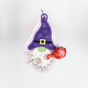 May include: Purple and white felt gnome ornament with a pointed hat, black belt, and a green beard. The gnome is holding a red lollipop.