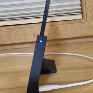 May include: A black tablet stand with a built-in charging port. The stand is angled, with a small blue light on the front. A white charging cable is connected to the stand. The stand is on a wooden surface.