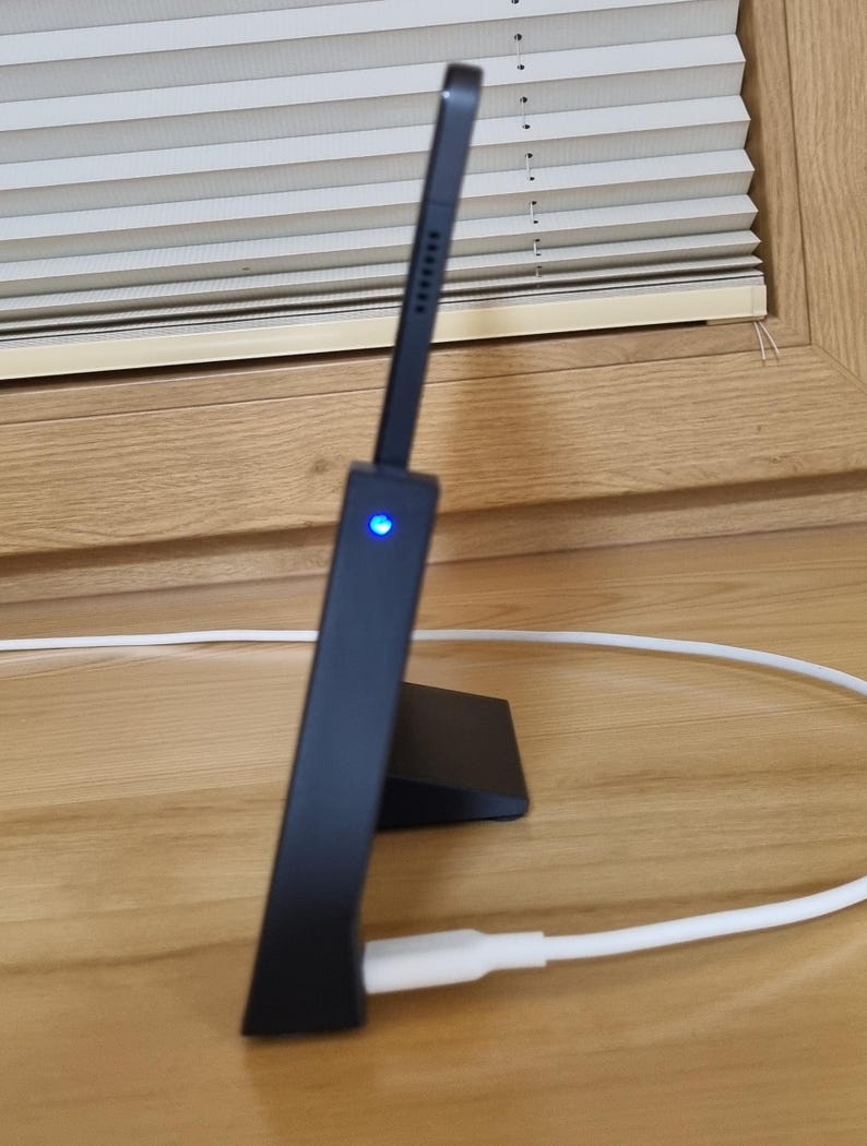 May include: A black tablet stand with a built-in charging port. The stand is angled, with a small blue light on the front. A white charging cable is connected to the stand. The stand is on a wooden surface.