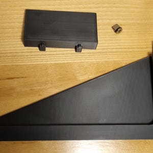 May include: Black wooden shelf bracket components, including a rectangular piece with two pegs, a triangular support, and a small metal fastener. The items are arranged on a light brown wooden surface.
